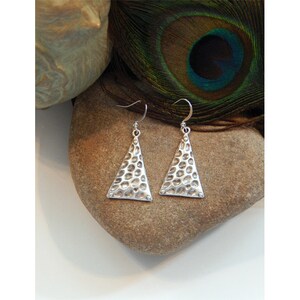 May include: A pair of silver triangle earrings with a hammered texture. The earrings are hanging from a stone surface.