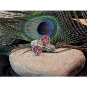 May include: Two round, pink, sparkly earrings on a gray stone. The earrings are set in silver.