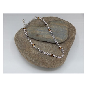 May include: A silver chain anklet with brown glass beads. The anklet is on a gray rock.
