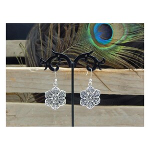 May include: A pair of silver-colored, filigree flower earrings with a detailed, openwork design. The earrings are displayed on a black stand against a backdrop of wood and a peacock feather.