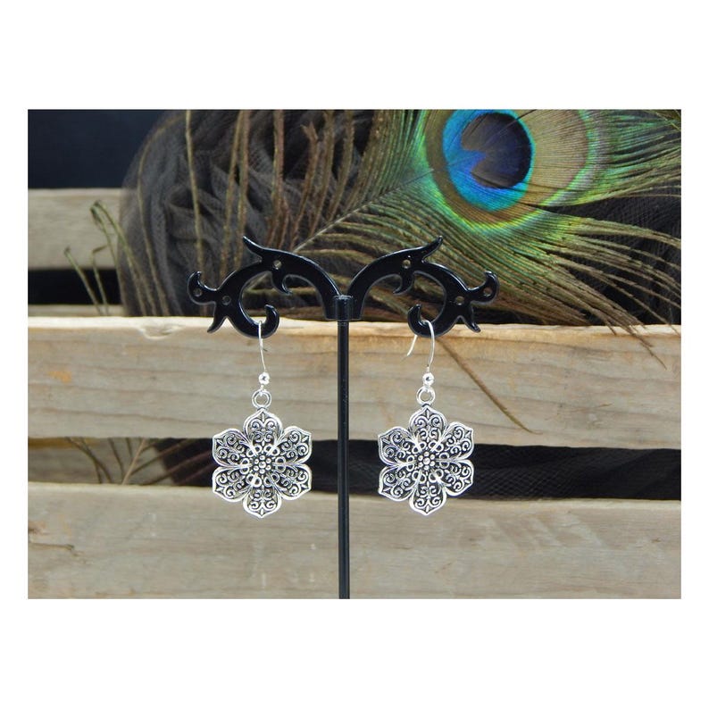 May include: A pair of silver-colored, filigree flower earrings with a detailed, openwork design. The earrings are displayed on a black stand against a backdrop of wood and a peacock feather.