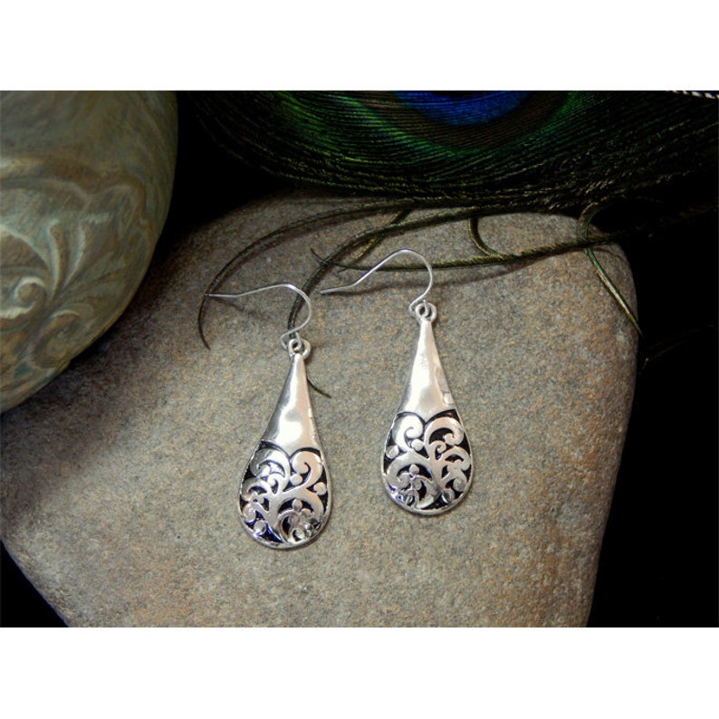 Puede incluir: Un par de pendientes de plata con forma de l&aacute;grima y un dise&ntilde;o filigrano intrincado. Los pendientes tienen un fondo negro y un contorno plateado.