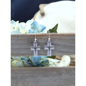 May include: A pair of silver cross earrings with an intricate design. The earrings are hanging from a wooden surface.