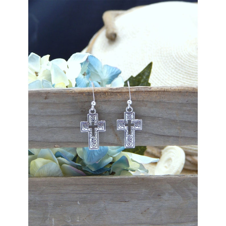 May include: A pair of silver cross earrings with an intricate design. The earrings are hanging from a wooden surface.