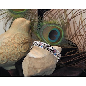 May include: A silver bracelet with a beaded design and colorful gemstones. The bracelet is displayed on a rock with peacock feathers in the background.