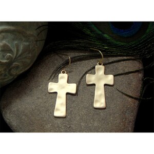 May include: A pair of gold-colored cross earrings with a hammered texture. The earrings hang from simple gold-tone ear wires. The crosses are displayed on a gray stone surface, with a dark background.