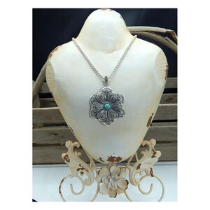 May include: A silver-toned pendant necklace with a large, ornate flower design. The pendant features a turquoise-colored stone in the center and is displayed on a white bust. The necklace is a statement piece, perfect for adding a touch of elegance.