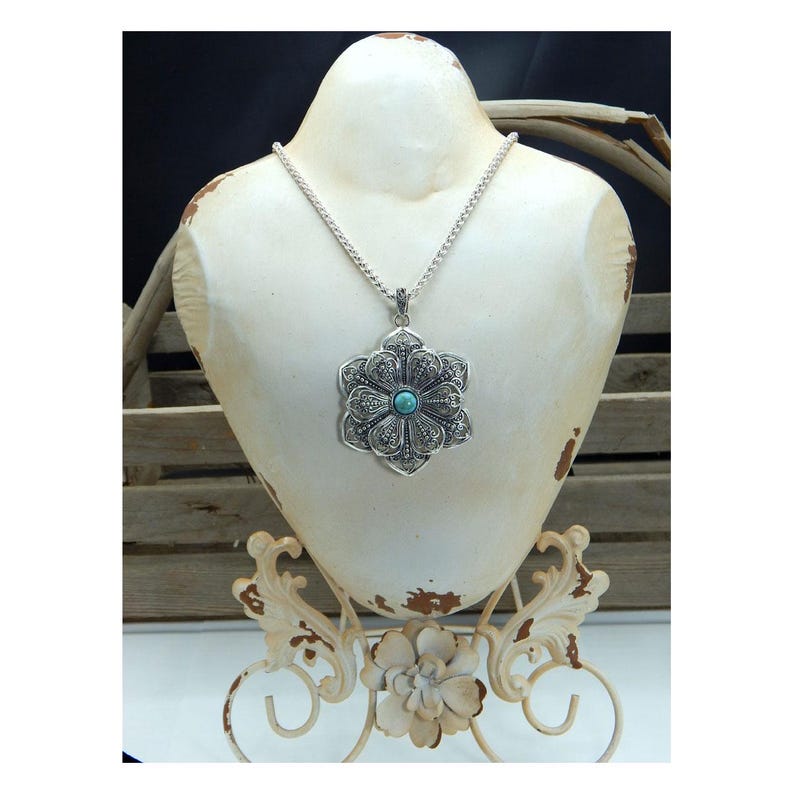May include: A silver-toned pendant necklace with a large, ornate flower design. The pendant features a turquoise-colored stone in the center and is displayed on a white bust. The necklace is a statement piece, perfect for adding a touch of elegance.