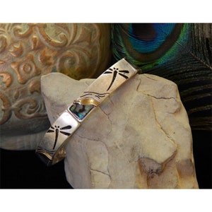 May include: Silver bracelet with a dragonfly design and abalone shell accents. The bracelet is on a gray rock.