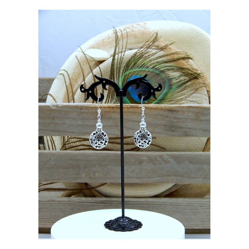 May include: A pair of silver-toned dangle earrings featuring circular charms with intricate openwork scroll patterns. The earrings hang from silver-toned hooks and are displayed on a black metal stand with a decorative, curved top design.