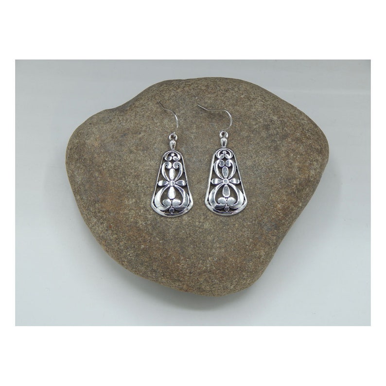 May include: A pair of silver dangle earrings with a delicate floral design. The earrings are hanging from a rock.