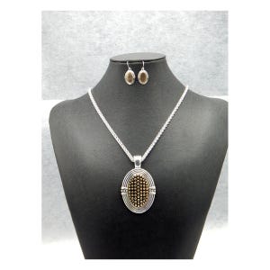 May include: A silver-toned necklace and matching earrings set. The pendant and earrings feature an oval design with a textured, gold-colored center and silver-toned borders. The necklace has a delicate chain.