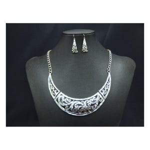 May include: A silver-toned necklace and earring set displayed on a black mannequin. The necklace is a crescent shape with intricate openwork scroll designs. The matching earrings are small, dangling pendants with similar detailing. The jewelry is set against a black background.
