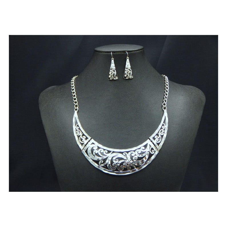 May include: A silver-toned necklace and earring set displayed on a black mannequin. The necklace is a crescent shape with intricate openwork scroll designs. The matching earrings are small, dangling pendants with similar detailing. The jewelry is set against a black background.