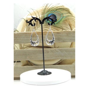 May include: Silver teardrop earrings with an open filigree design, displayed on a black stand. The earrings have a detailed, openwork pattern with dark accents. The stand is on a white, round base, with a wooden background.