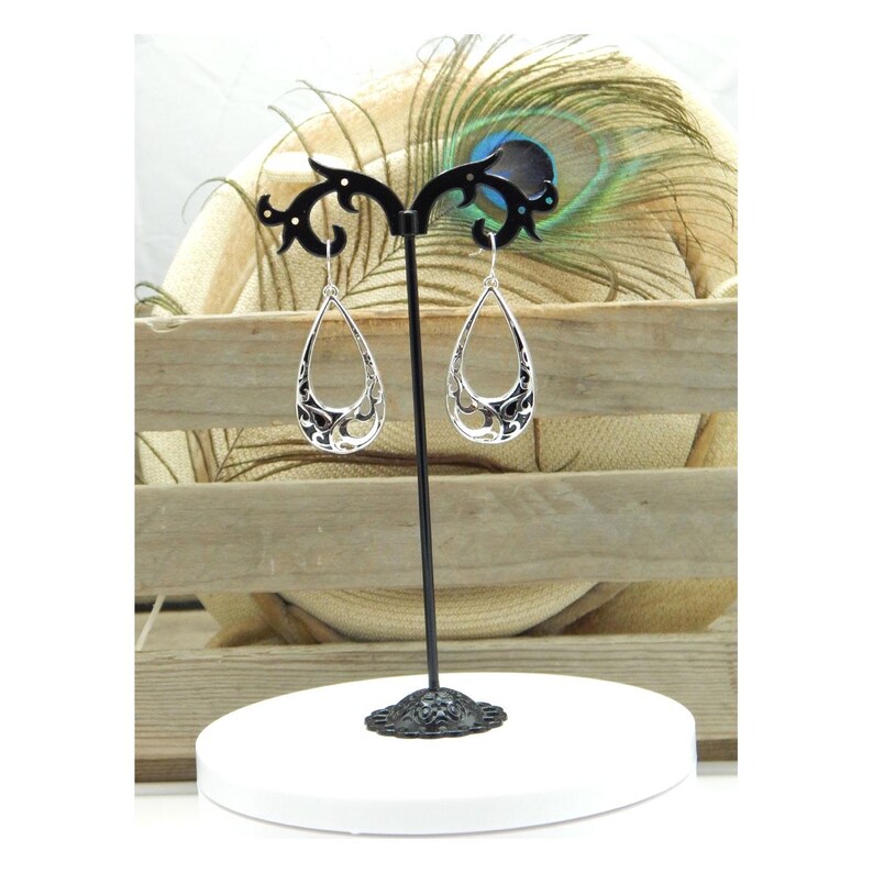 May include: Silver teardrop earrings with an open filigree design, displayed on a black stand. The earrings have a detailed, openwork pattern with dark accents. The stand is on a white, round base, with a wooden background.