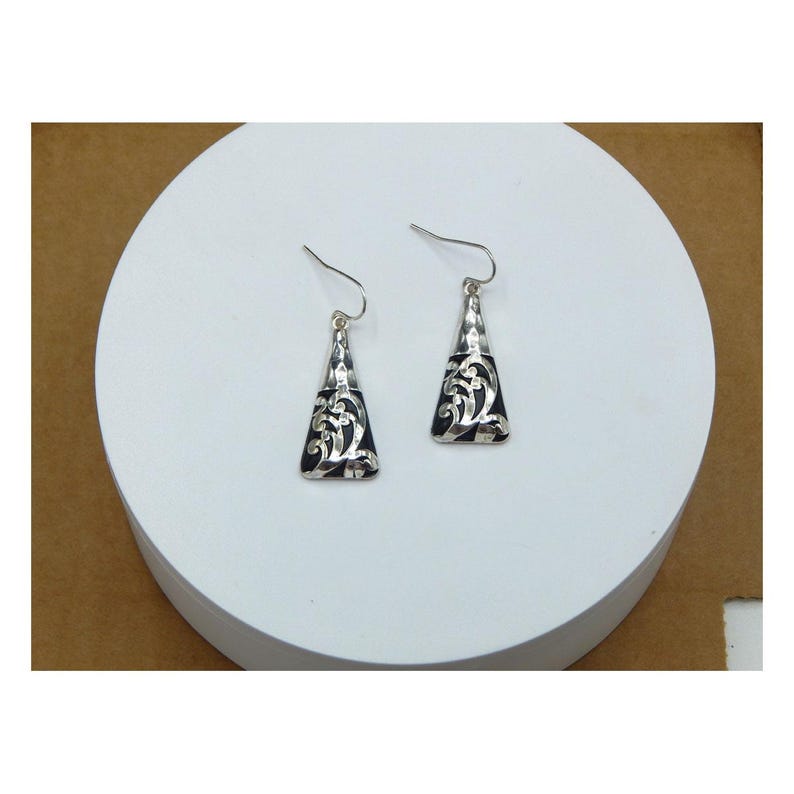 May include: A pair of silver-toned, triangular dangle earrings. The earrings have a black background with a silver filigree design. The earrings are displayed on a white circular surface.
