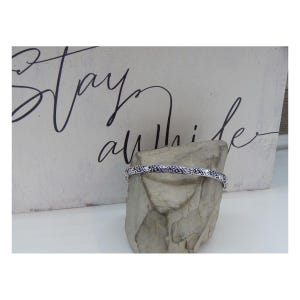 May include: A silver bangle bracelet with an intricate, detailed design. The bracelet is displayed on a small, grey rock.