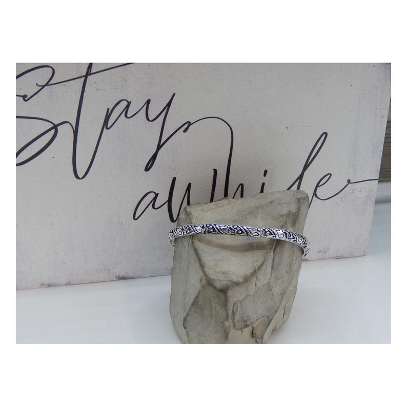 May include: A silver bangle bracelet with an intricate, detailed design. The bracelet is displayed on a small, grey rock.