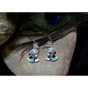 May include: A pair of silver cat earrings with colorful enamel details. The cats are sitting with their tails curled around their legs. The enamel is in shades of blue, pink, yellow, and black.