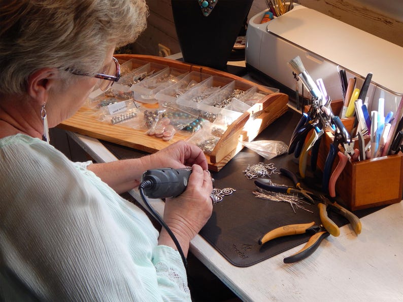 May include: A person working on jewelry, using a rotary tool. The workspace includes a wooden organizer with jewelry components, tools, and a variety of pliers. The scene suggests a jewelry-making studio.