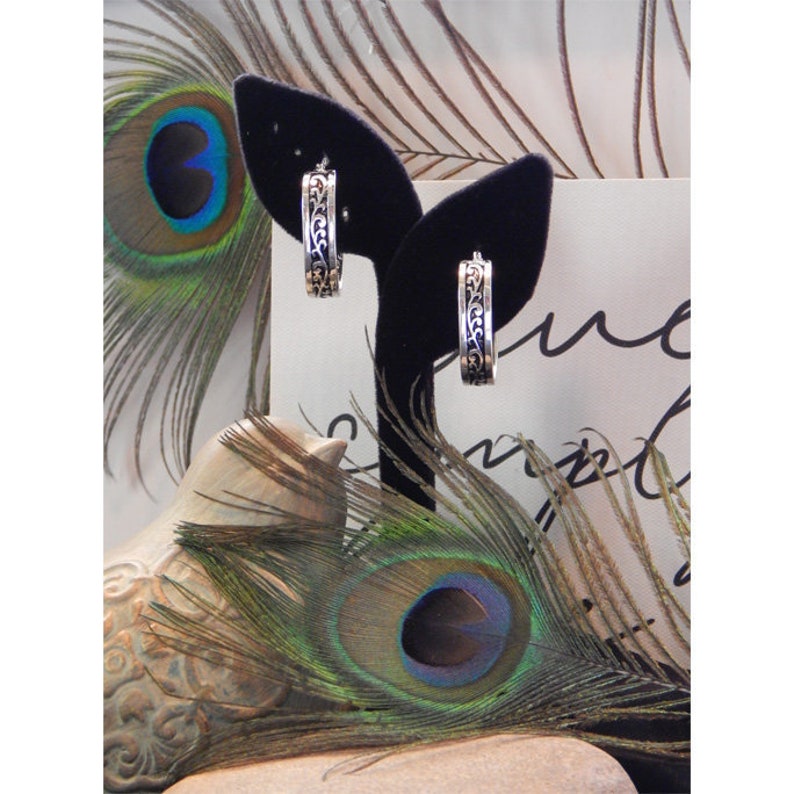 May include: A pair of silver hoop earrings with an intricate floral design. The earrings are displayed on a black velvet stand against a background of peacock feathers.