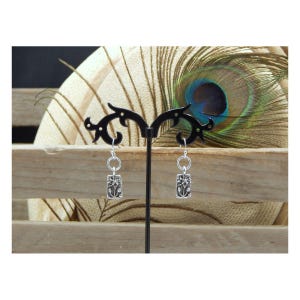 May include: A pair of silver earrings with a rectangular design featuring a floral pattern. The earrings are displayed on a black stand against a backdrop of a peacock feather and wooden planks.