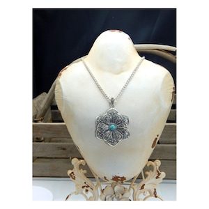 May include: A silver-toned necklace with a large, ornate flower pendant. The pendant features a turquoise-colored stone in the center and intricate detailing. The necklace is displayed on a white mannequin bust.