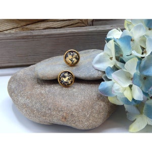 May include: A pair of gold stud earrings with black resin and gold leaf accents. The earrings are displayed on a gray rock.