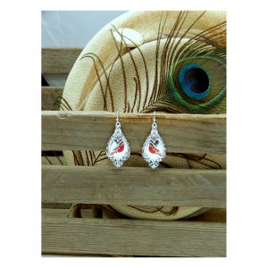 May include: A pair of silver-toned, teardrop-shaped earrings. Each earring features a red cardinal bird design within a decorative frame. The earrings hang from a simple hook. The background includes a wooden crate and a peacock feather.