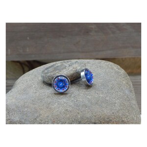 May include: A pair of round stud earrings with a blue and red floral design. The earrings have a silver-colored metal setting and are displayed on a gray stone. The design features a central blue circle with red and white accents.