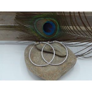 May include: A pair of silver hoop earrings with a textured finish. The earrings are large and circular, and they are lying on a gray rock.