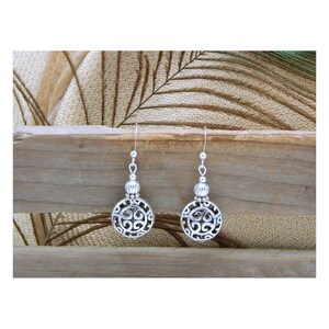 May include: A pair of silver-toned drop earrings featuring circular pendants with an intricate openwork scroll pattern. Each earring is suspended from a simple hook wire and includes a small, ribbed spherical bead accent above the main disc.