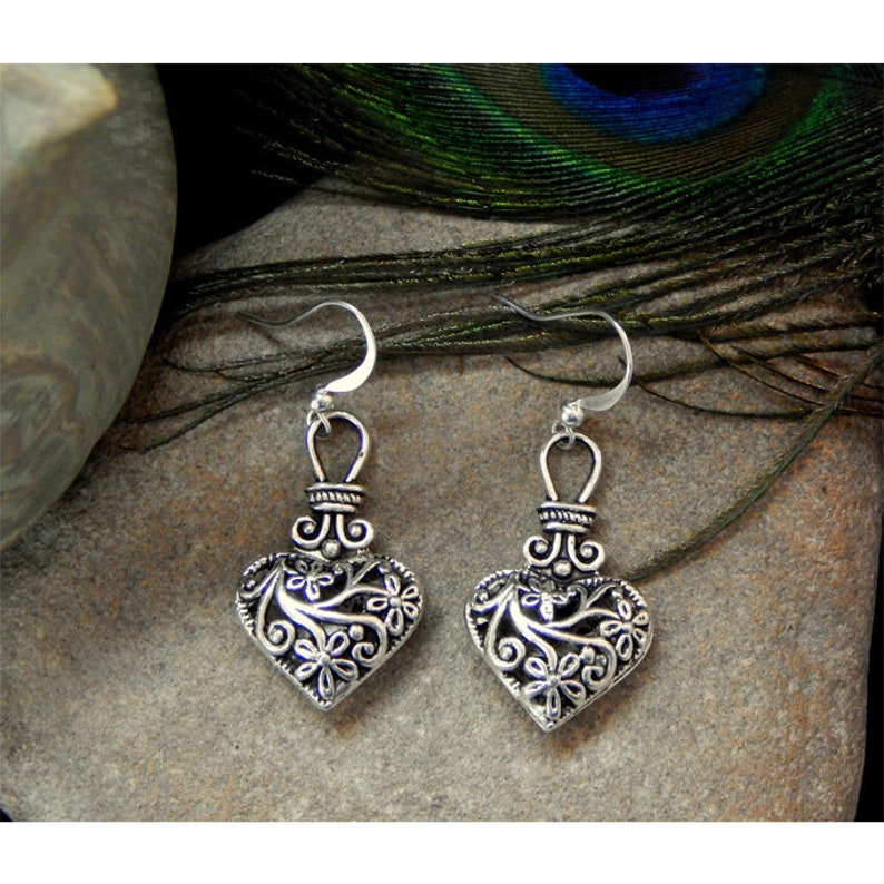 May include: A pair of silver heart-shaped earrings with intricate floral designs. The earrings have a dangling design with a small loop at the top for attaching to an earring hook.