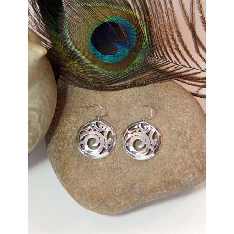 May include: Silver dangle earrings with a circular design featuring a swirl pattern. The earrings are on a gray stone surface.