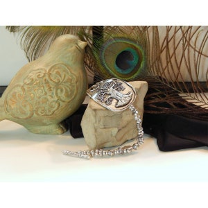 May include: A silver bracelet with a tree of life charm. The charm is embossed with a detailed tree design. The bracelet is made of silver beads and has a clasp closure.