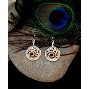 May include: A pair of silver and gold earrings with a circular design. The earrings feature a silver outer ring with an intricate pattern and a gold-colored swirl design in the center. The earrings are displayed on a gray stone, with a peacock feather in the background.