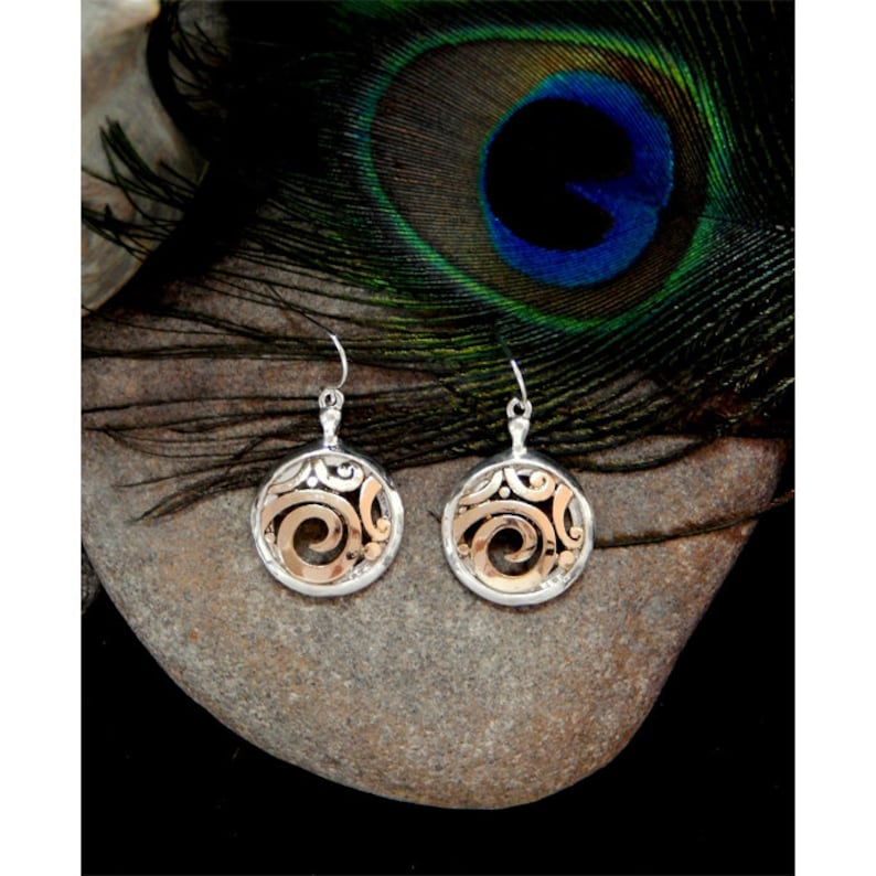 May include: A pair of silver and gold earrings with a circular design. The earrings feature a silver outer ring with an intricate pattern and a gold-colored swirl design in the center. The earrings are displayed on a gray stone, with a peacock feather in the background.
