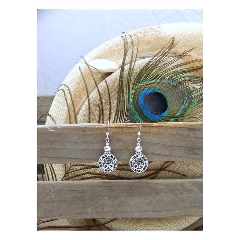 May include: A pair of silver-toned dangle earrings featuring circular charms with intricate openwork scroll patterns. Each earring is suspended from a simple hook wire and includes a small, textured silver bead accent above the circular charm.