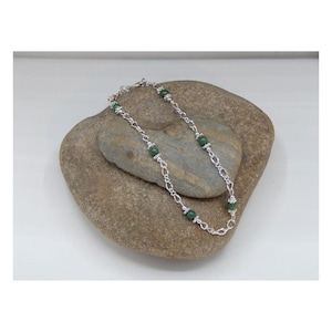 May include: A silver-toned necklace with green beads and a delicate chain design. The necklace is displayed on a gray stone, showcasing its elegant simplicity. The necklace is a piece of jewelry.