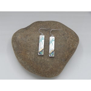 May include: A pair of silver dangle earrings with a dragonfly design. The earrings have a rectangular shape and are decorated with blue and green enamel. The dragonfly is outlined in silver and has a light blue body.