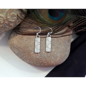 May include: Silver dangle earrings with a geometric pattern. The earrings are hanging from a stone surface.