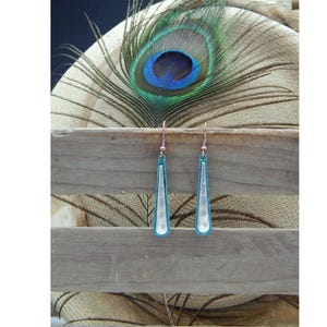 May include: A pair of dangle earrings with a teardrop-shaped silver pendant and a turquoise blue border. The earrings are hanging from a wooden surface.
