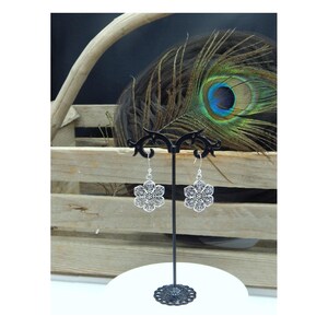 May include: A pair of silver-colored, openwork flower earrings on a black display stand. The earrings have a detailed, symmetrical design. The background includes a wooden crate, a peacock feather, and a white surface.