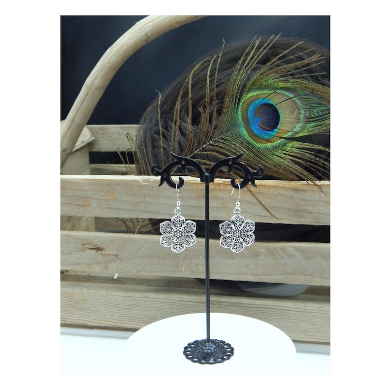 May include: A pair of silver-colored, openwork flower earrings on a black display stand. The earrings have a detailed, symmetrical design. The background includes a wooden crate, a peacock feather, and a white surface.