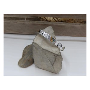 May include: Silver bangle bracelet with a tree design and colorful inlay. The bracelet is on a gray rock.