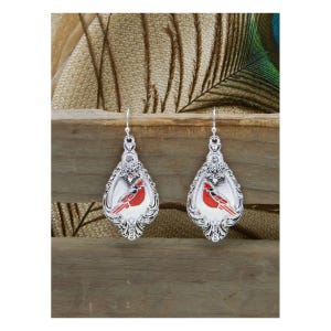 May include: Silver-toned drop earrings with a red cardinal bird design. The earrings have a decorative, ornate border and a hook closure. The cardinal is depicted in a vibrant red color, set against a white background.