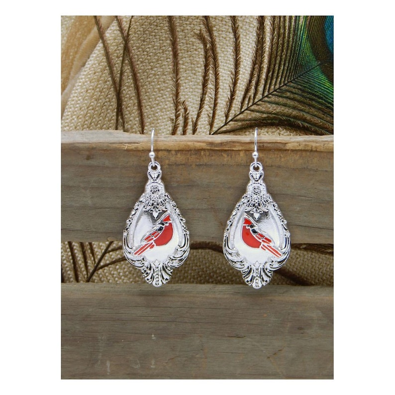 May include: Silver-toned drop earrings with a red cardinal bird design. The earrings have a decorative, ornate border and a hook closure. The cardinal is depicted in a vibrant red color, set against a white background.