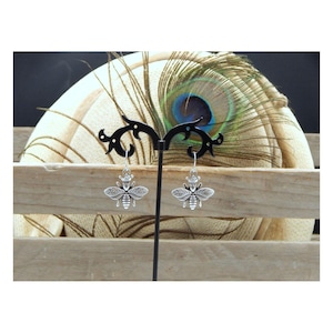 May include: A pair of silver-colored bee earrings. Each earring features a detailed bee design with a crown on its head. The earrings are displayed on a black earring stand against a backdrop of a peacock feather and wooden slats.