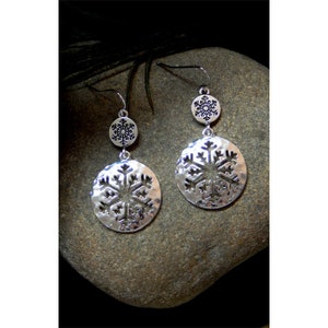 May include: Silver-toned dangle earrings featuring a snowflake design. Each earring has a small circular top with a snowflake and a larger circular pendant with a cut-out snowflake design. The earrings are displayed on a gray stone.